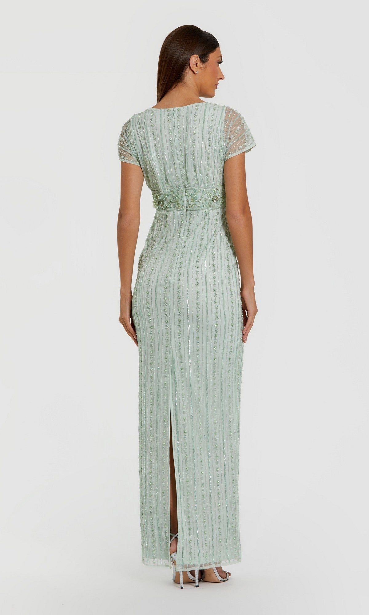 A woman stands with her back to the camera in the Mac Duggal 94147 Long Formal Dress—a floor-length, light green gown adorned with beaded pearl embellishments, subtle vertical detailing, short sleeves, and a back slit—set against a plain white background.